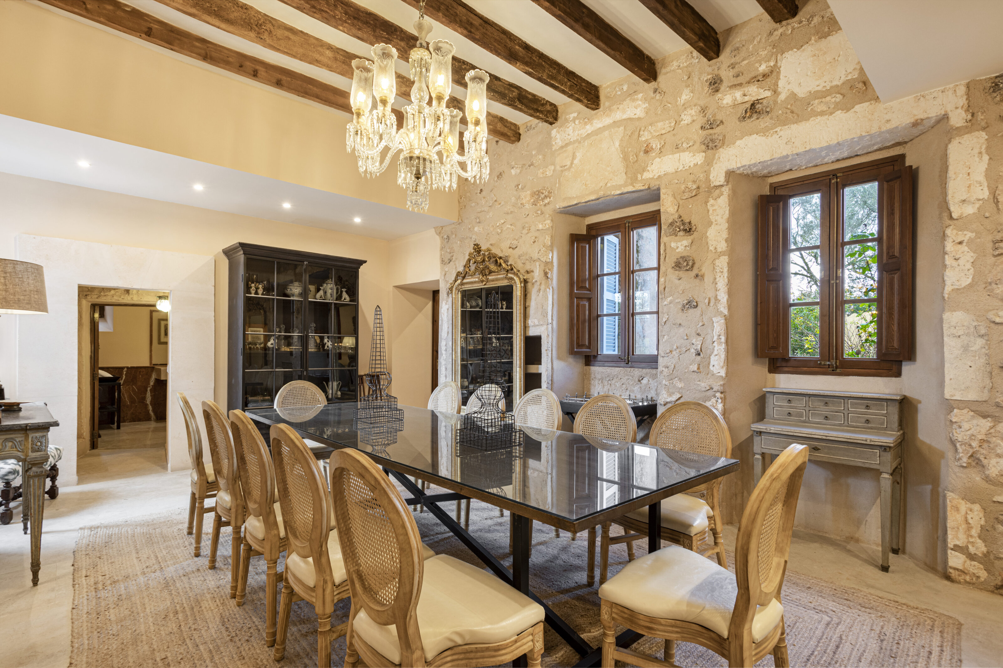 A dining room with a long table, cane chairs, a crystal chandelier and sunlit stone walls